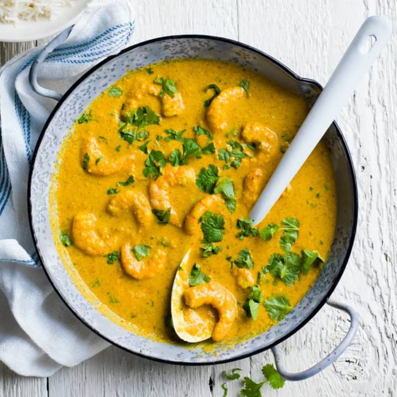 A pan of vibrant yellow Indian shrimp curry with cilantro, a white spoon, and a napkin.