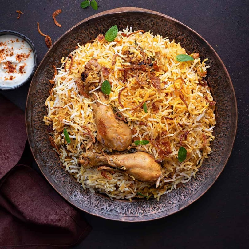 Chicken Biryani with two drumsticks, fried onions, and mint, served with raita in a bowl.