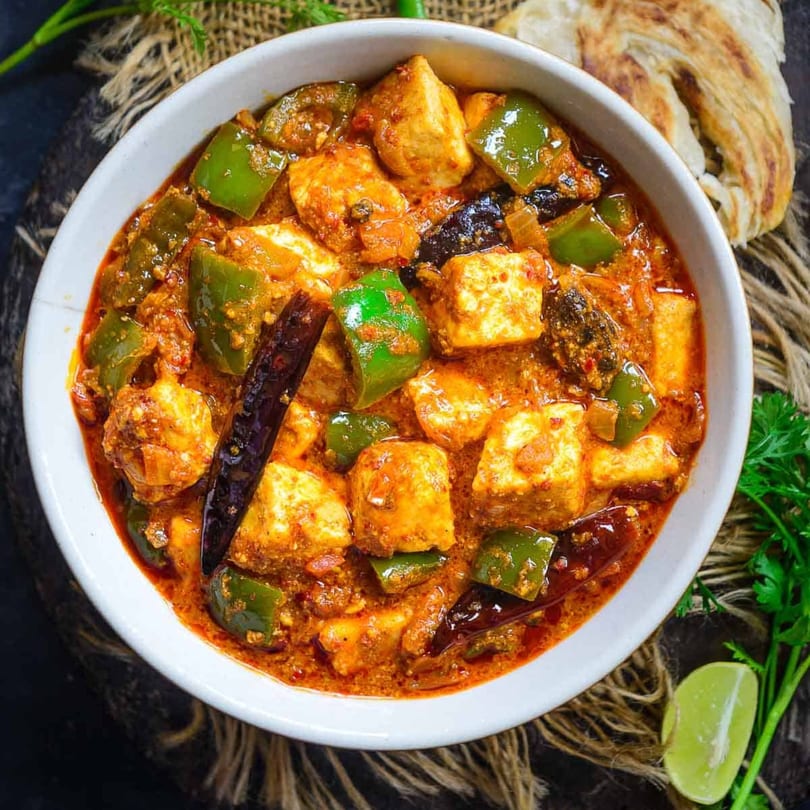 A bowl of Indian Kadai Paneer with bell peppers, spices, and a side of paratha and lime.