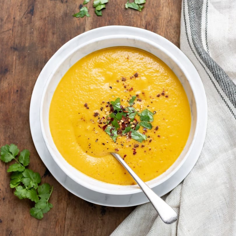 Overhead view of a bowl of vibrant orange Indian soup, garnished with cilantro and chili flakes.
