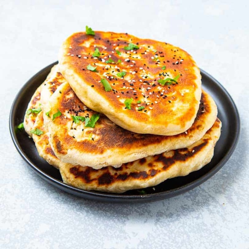 Stack of golden-brown Indian naan bread, garnished with sesame seeds and fresh herbs on a black plate.